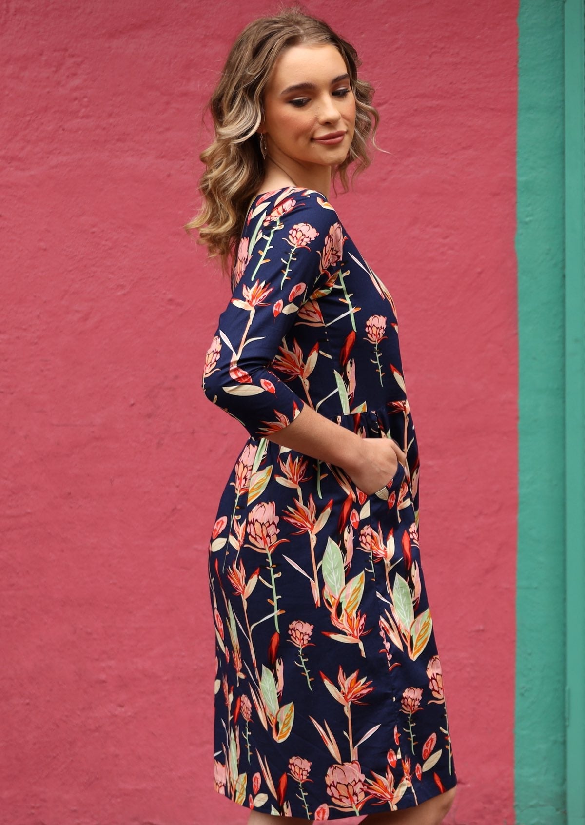 Model wears cotton pink, green and blue native botanic print knee length dress with her hands in pockets