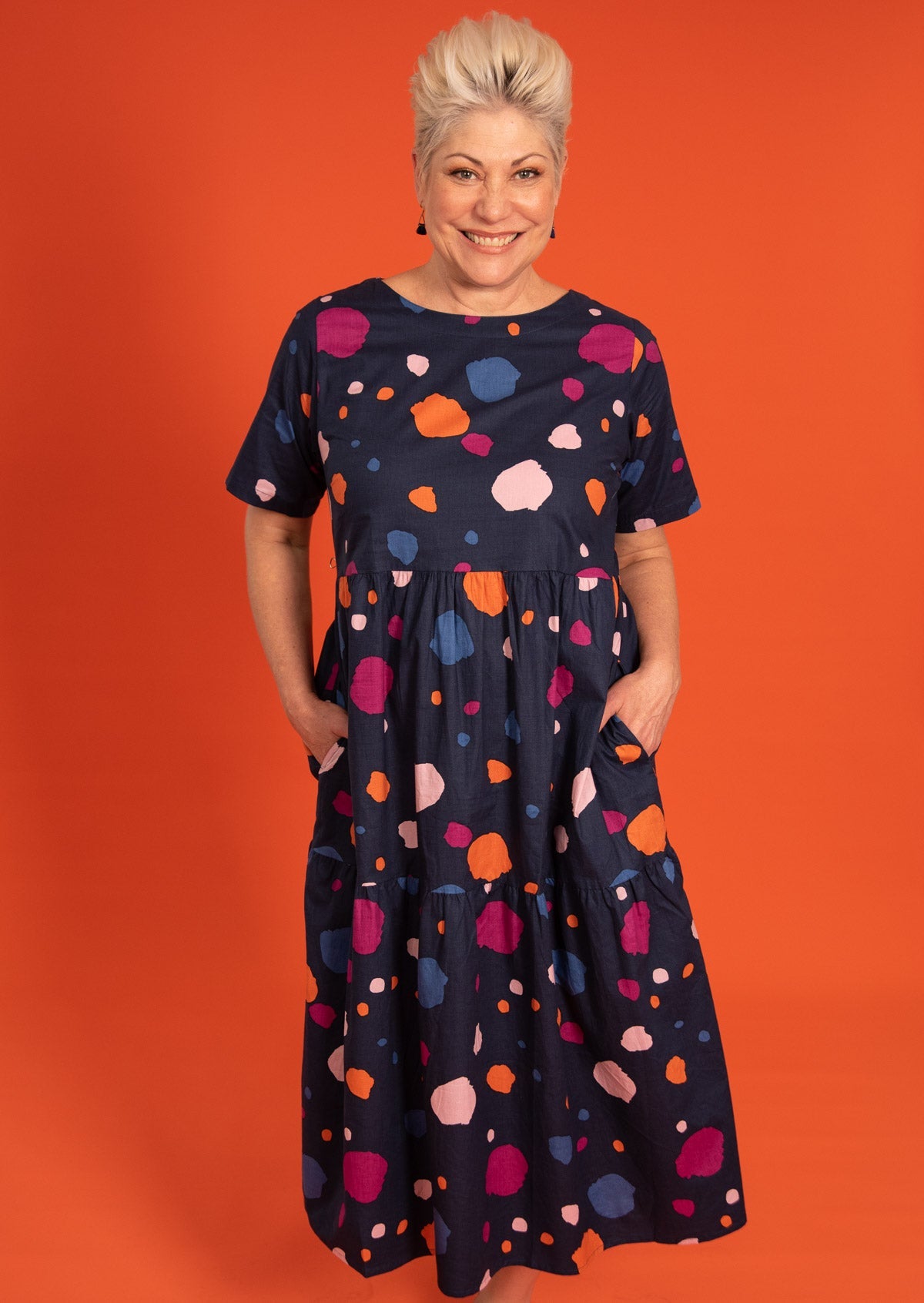 Model wears 100% cotton midlength navy dress in bold colorful print with round neckline and hands in pocket.