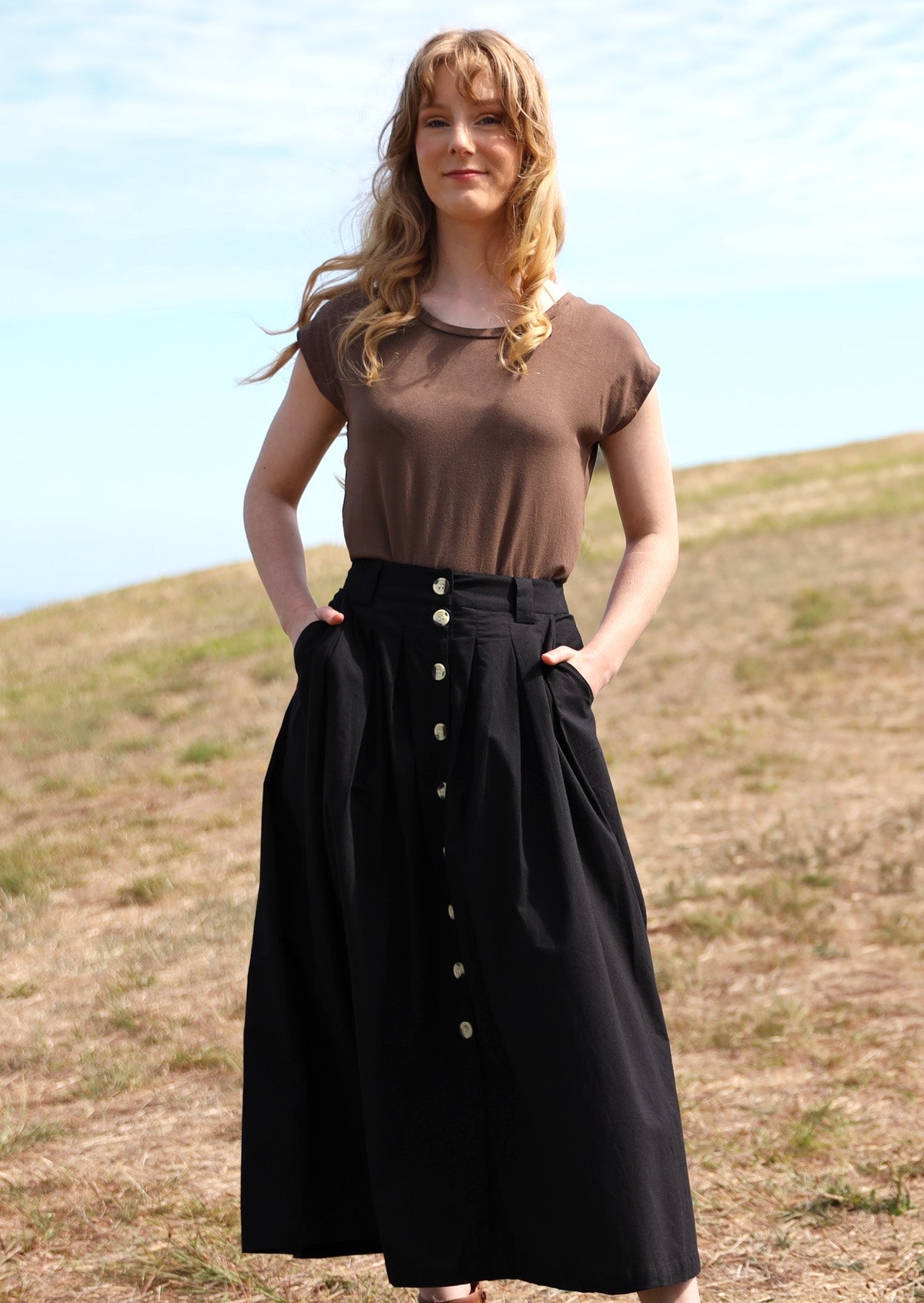 Woman with red hair wearing Cora Skirt full front button down front belt loops box pleats maxi skirt with side pockets 100% cotton black with light brown t-shirt 