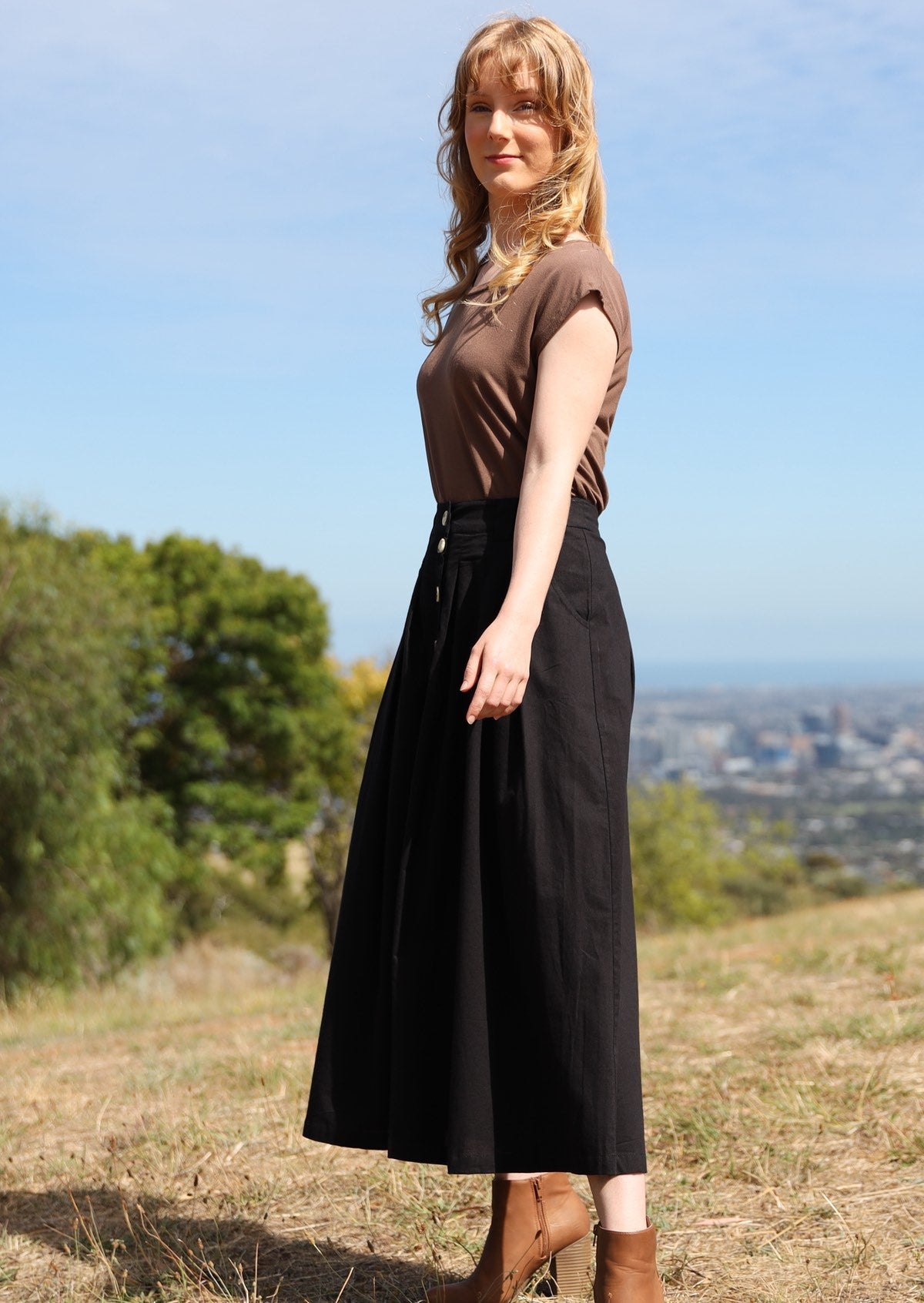 Woman stands side on in a cotton midi length black skirt, paired with a brown t-shirt and tan ankle boots 