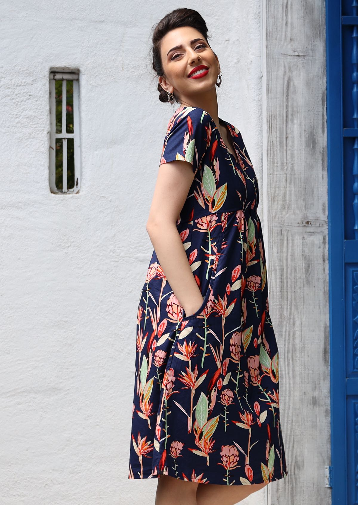 Model wears colorful native pink and green floral printed 100% cotton knee length dress with hands in pocket.