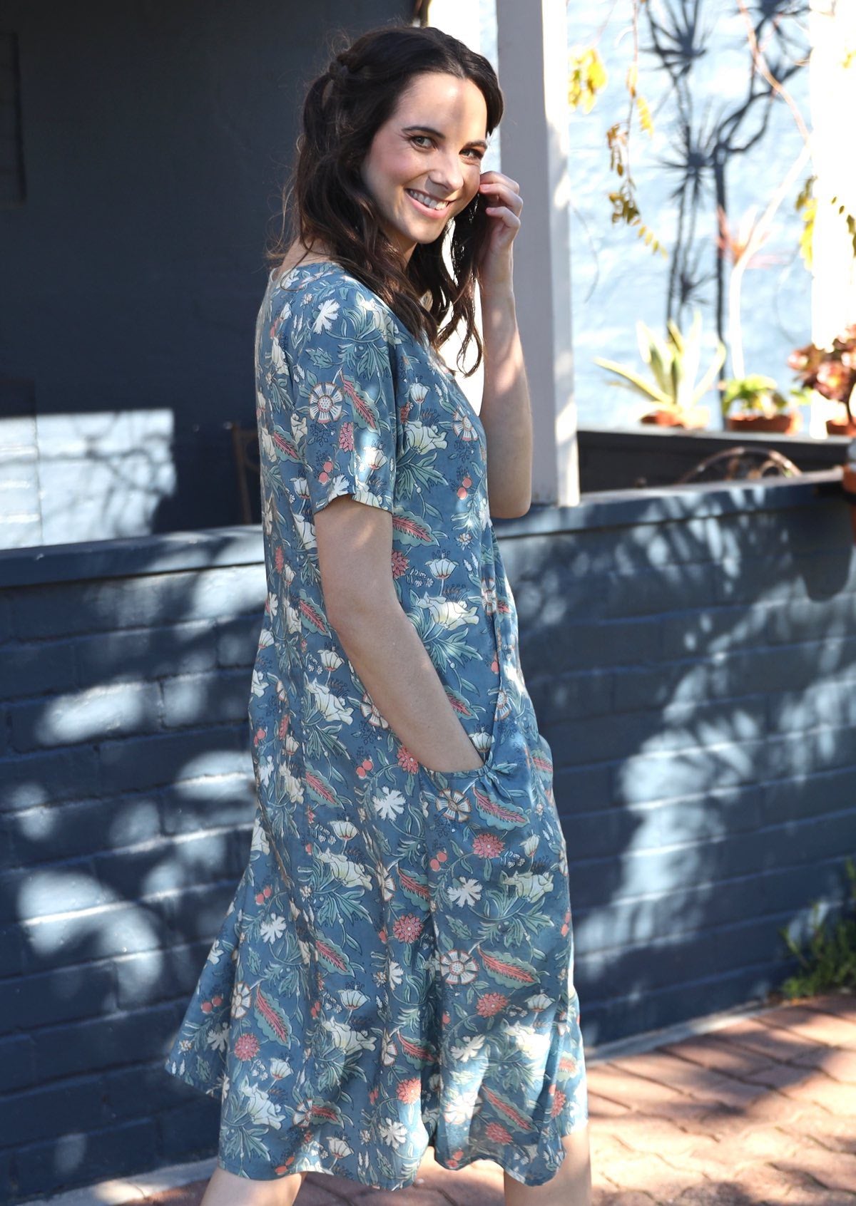 Woman walking away in dappled sunlight, wearing a cotton floral print dress with hand deep in pocket