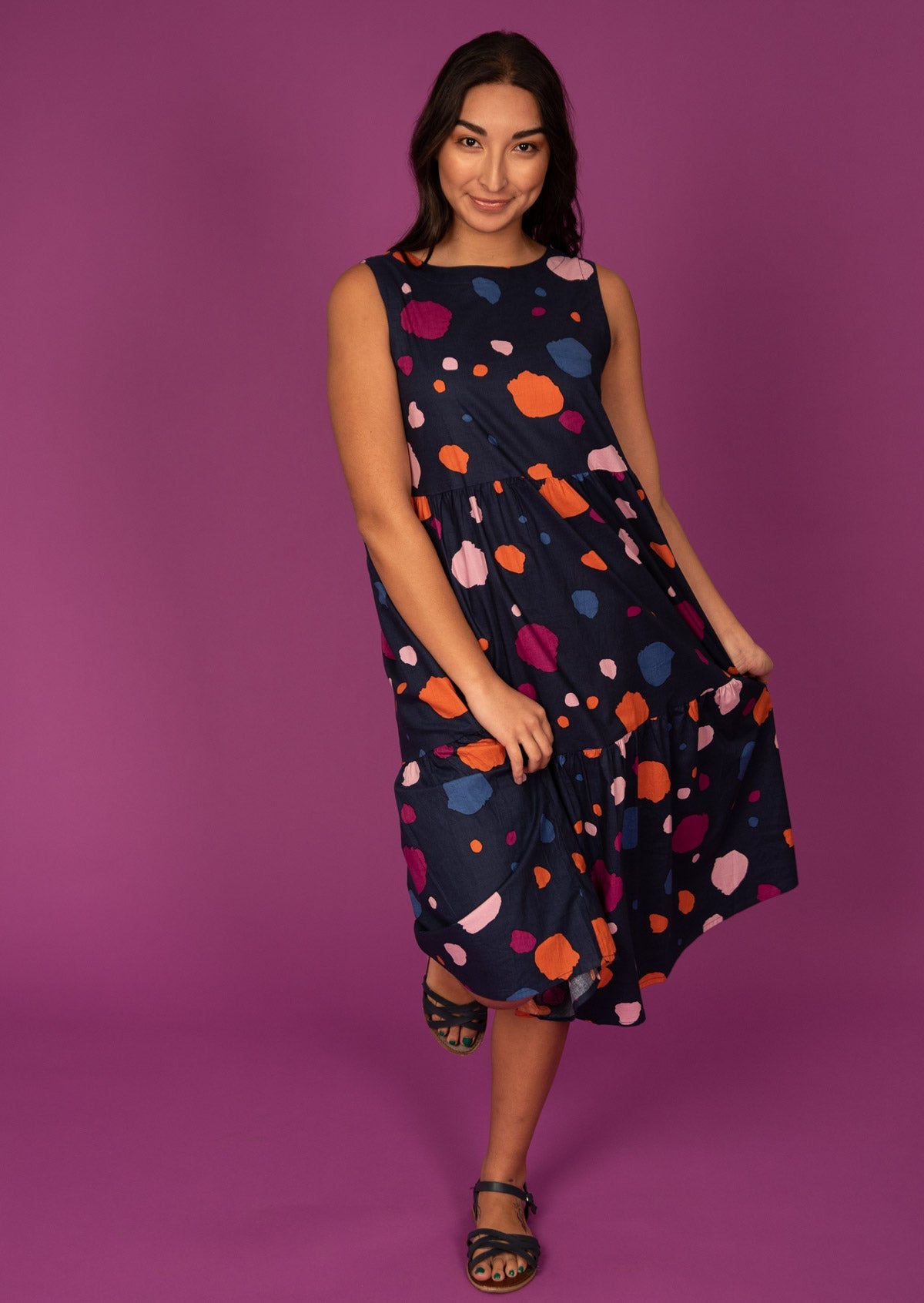 Model standing in front of pink background wears loose fitted sleeveless cotton dress with pockets.

