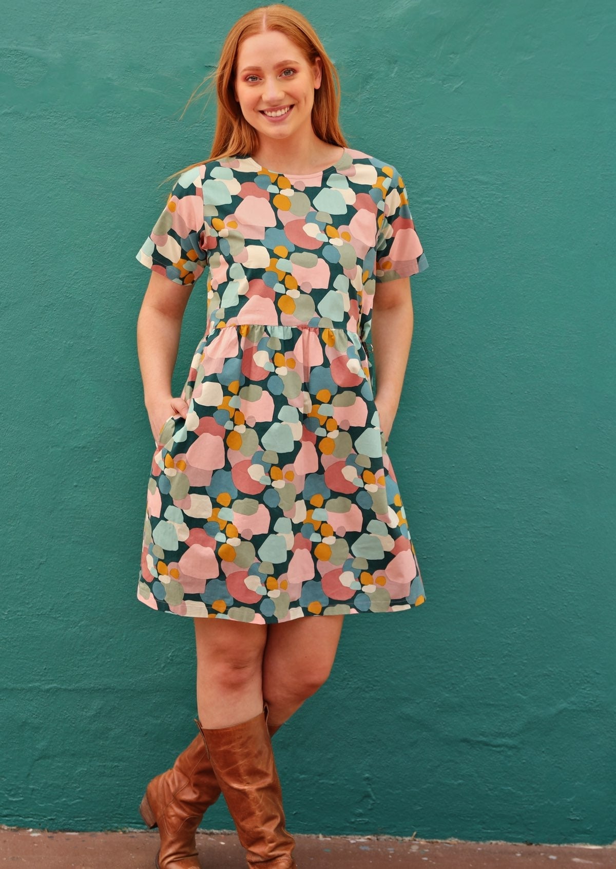 Woman wears above knee length cotton dress in colorful pastel spotty print, with leather boots.