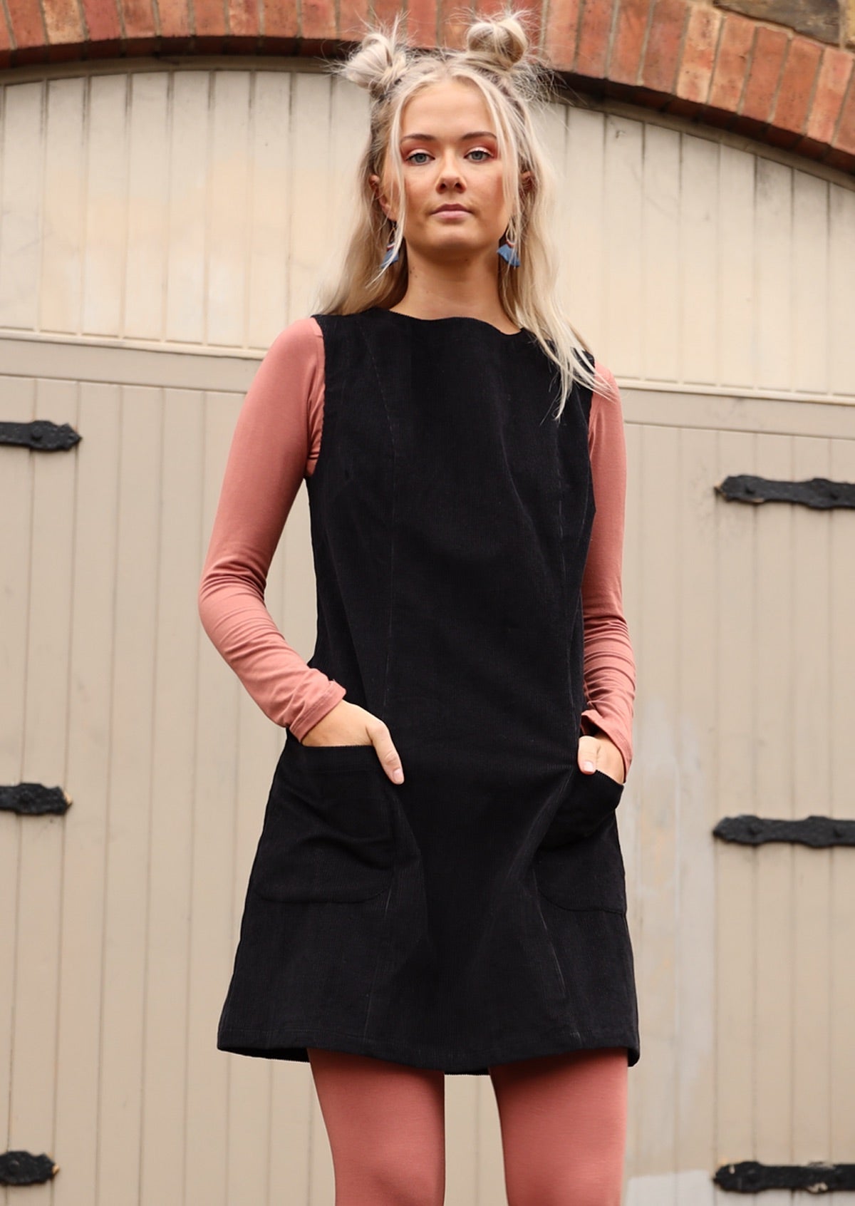 Model poses in sleeveless black corduroy above knee tunic with hands in front pockets