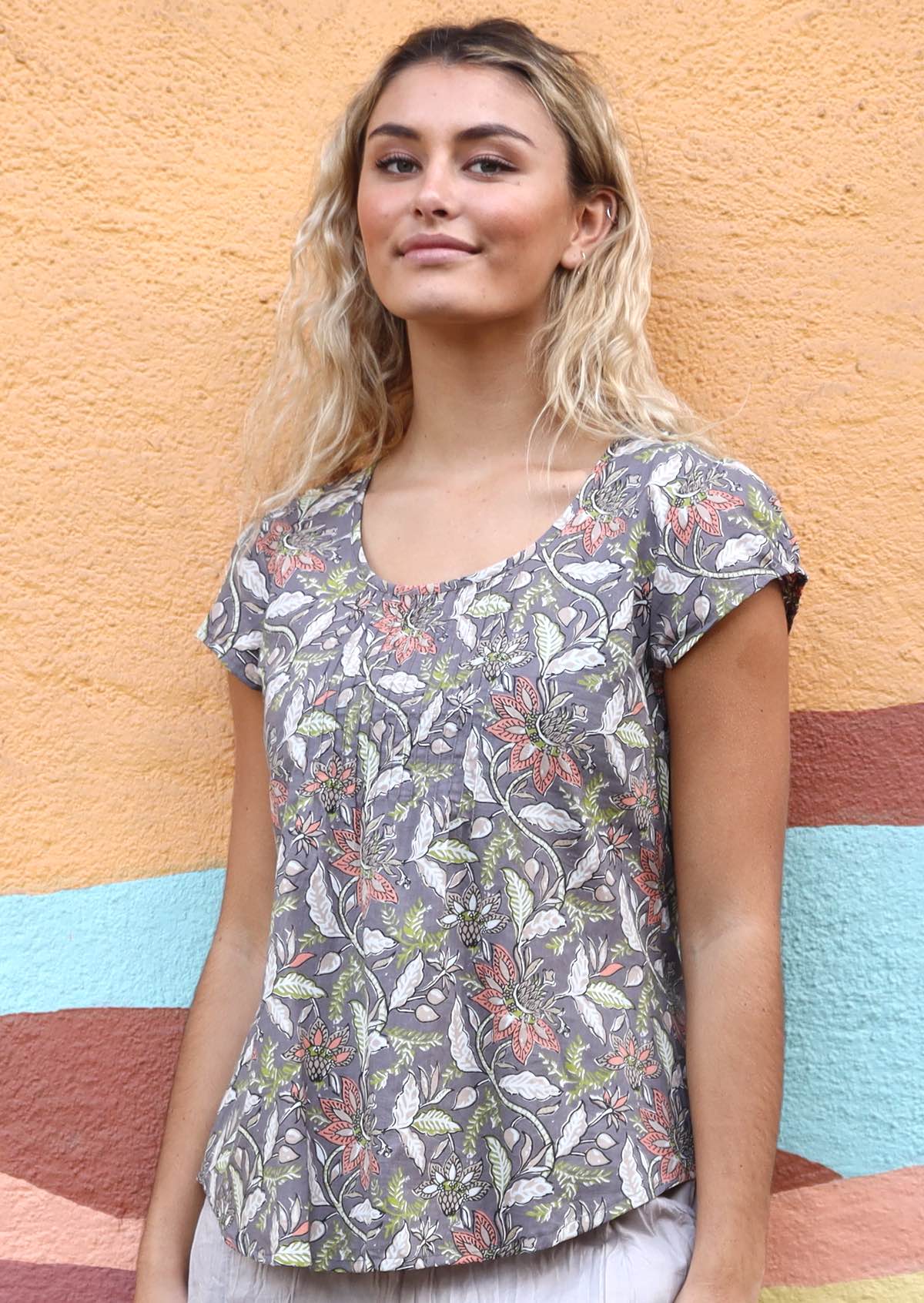 Woman leans against wall in taupe based floral print cap sleeve cotton top